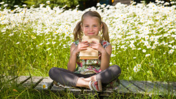 Tyttö istuu ulkona kirja kädessä. Kesäinen maisema taustalla.